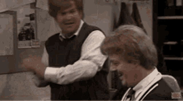 two men are standing next to each other in a room . one of the men is wearing a vest .