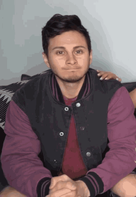 a man wearing a purple shirt and a black jacket is sitting on a couch