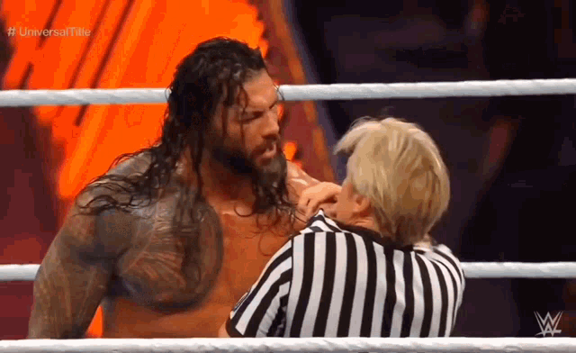 roman reigns is talking to a referee in a wrestling ring during a match .