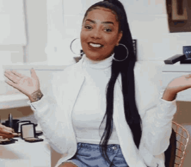 a woman wearing a white jacket and denim shorts is sitting in front of a makeup table .