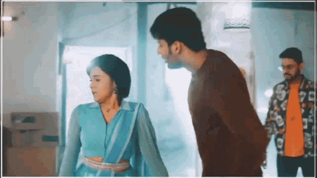 a man and a woman are standing next to each other in a room . the woman is wearing a blue saree .