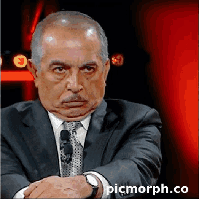 a man in a suit and tie is sitting with his hands folded in front of a red background with picmorph.co written below him