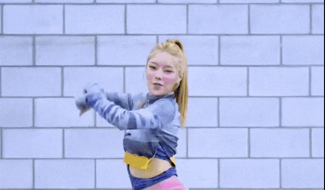a woman in a blue sweater is dancing in front of a white brick wall