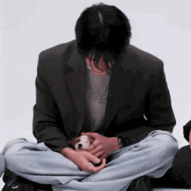 a man in a black jacket is sitting on the floor with his legs crossed holding a small white dog .