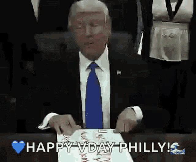 a man in a suit and tie is sitting at a table holding a piece of paper that says happy vday philly .
