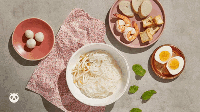a bowl of noodles sits on a table next to plates of shrimp and eggs