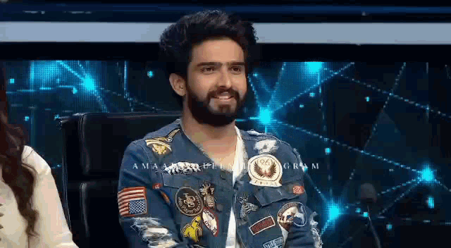 a man with a beard is sitting in a chair wearing a denim jacket with patches .