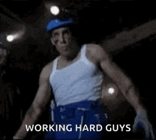 a man in a white tank top and blue shorts is standing in a dark room and saying `` working hard guys '' .