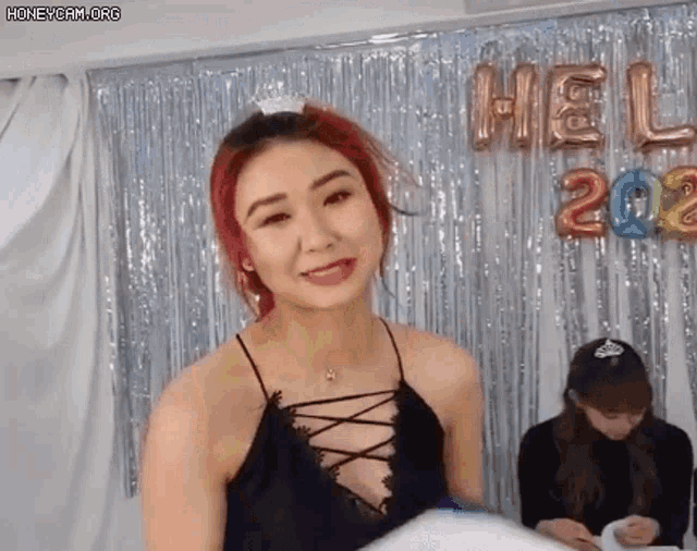 a woman in a black lace top is standing in front of a curtain with balloons that say hello 2022 .