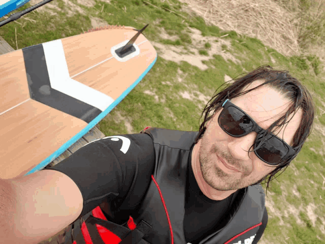 a man wearing sunglasses takes a selfie with a surfboard in the background