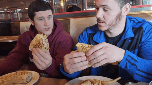a man in a blue nike jacket is eating a sandwich with another man