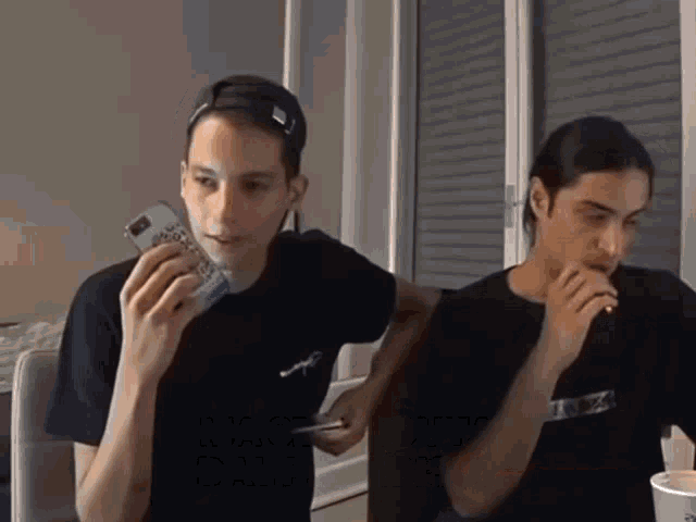 a man in a black t-shirt holds a cell phone in his right hand while another man looks on
