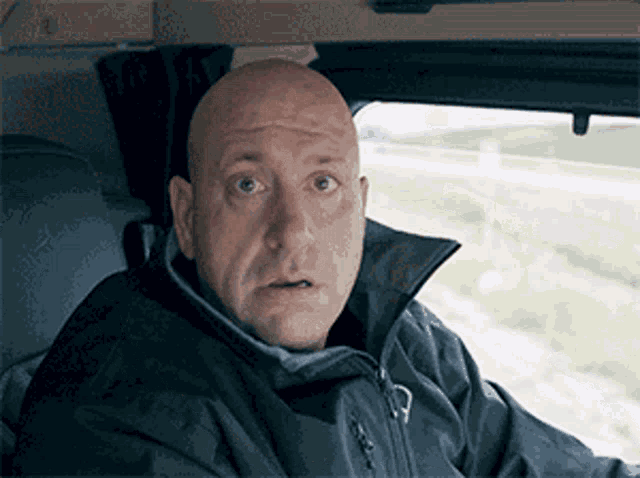 a bald man wearing a black jacket looks surprised
