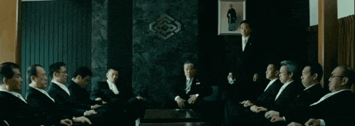 a group of men in suits are sitting around a table with a picture of a man on the wall behind them