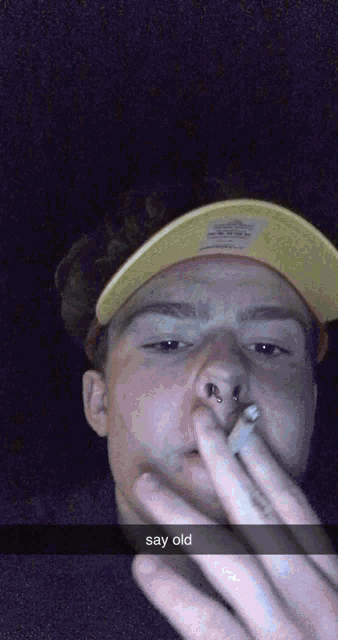 a man wearing a yellow hat is smoking a cigarette with the caption say old