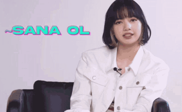 a woman in a white jacket is sitting in a chair with sana ol written on a wall behind her