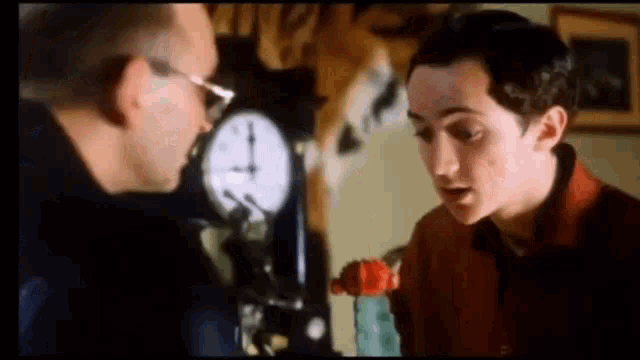 two men are talking in front of a clock .