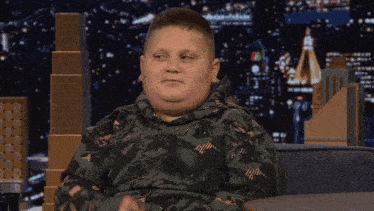 a young boy wearing a camouflage sweatshirt is sitting in front of a city at night