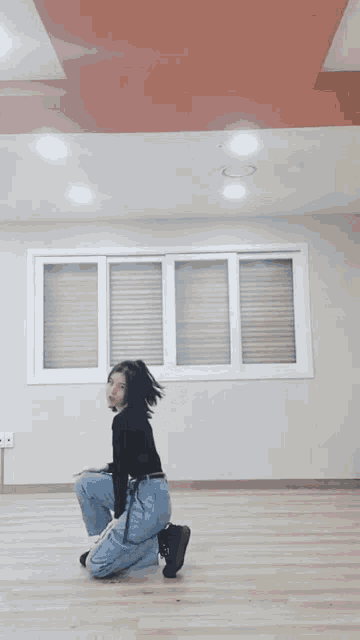 a woman in a black top and blue jeans is kneeling on the floor in front of a window