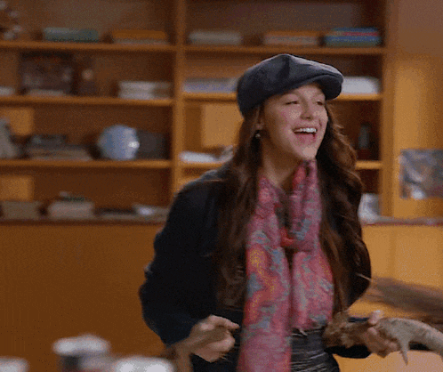 a woman wearing a scarf and a hat smiles while holding a piece of wood