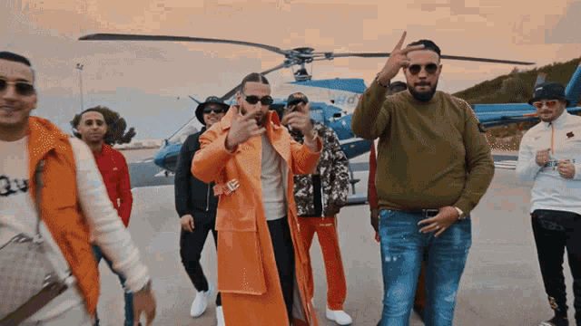 a group of men are standing in front of a helicopter and one of them is wearing an orange coat