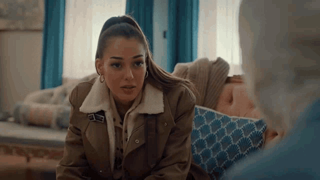 a woman in a brown jacket sits on a couch talking to another woman