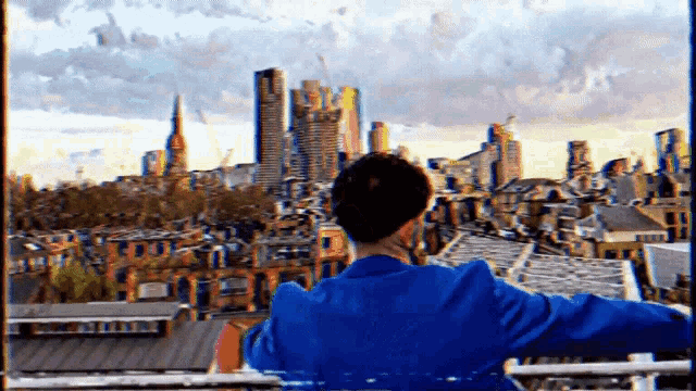 a man in a blue jacket stands on a balcony overlooking a city skyline