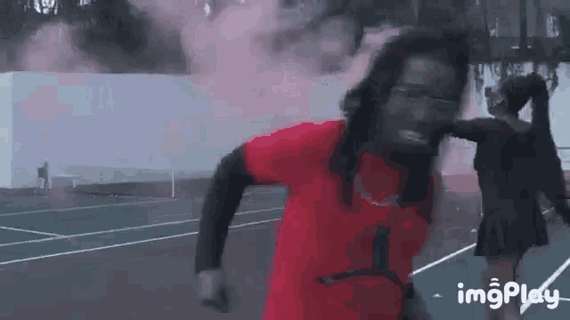 a man in a red shirt is running on a tennis court while a woman in a black dress stands behind him .