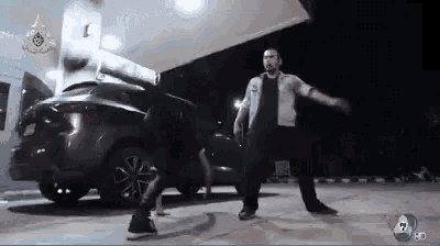 two men are dancing in front of a car in a parking lot at night .