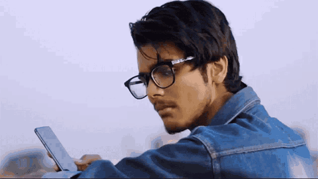 a young man wearing glasses and a denim jacket looks at his phone