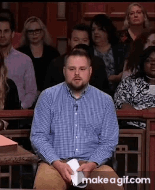 a man in a blue plaid shirt is sitting in front of a crowd in a courtroom .