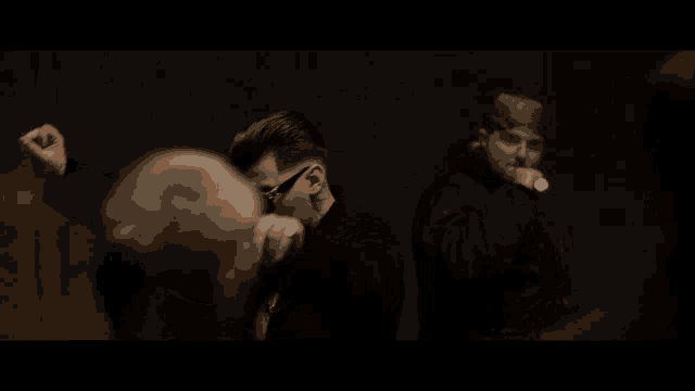two men standing next to each other in a dark room with one wearing sunglasses