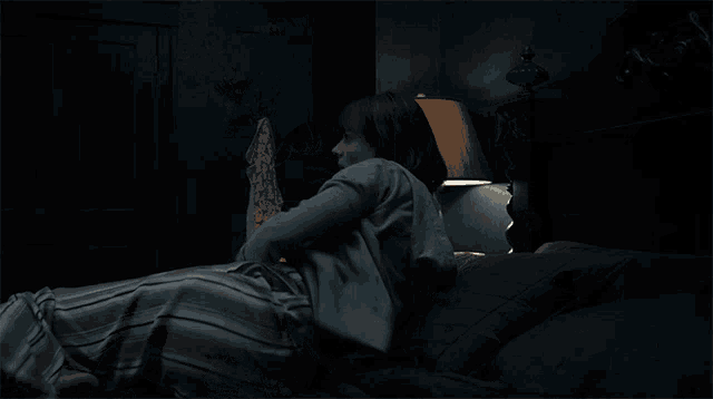 a woman sits on a bed in a dark room with a lamp