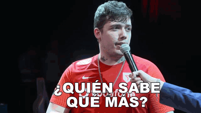 a man in a red shirt is talking into a microphone and the words quien sabe que mas are above him
