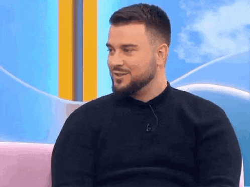 a man with a beard is wearing a black sweater and sitting on a pink couch .