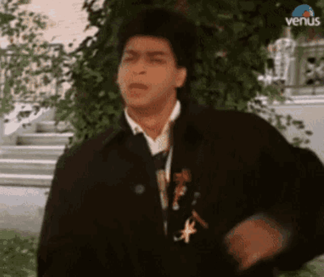 a man in a black jacket and tie is standing in front of a tree and stairs .