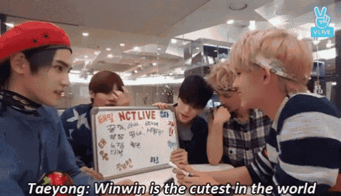 a group of young men are sitting around a table with a white board that says nctlive on it .