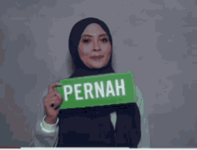 a woman in a blue hijab holds a green sign that says pernah