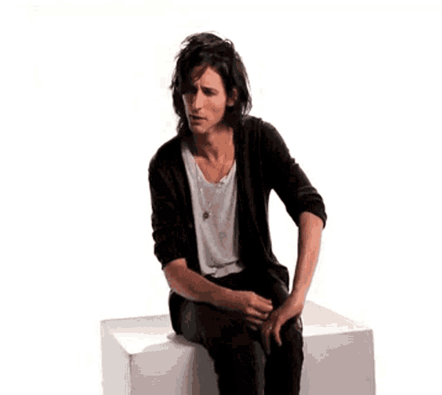 a young man is sitting on a white box with his hands on his knees .