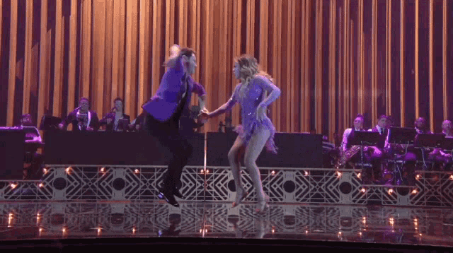 a man and a woman are dancing on a stage with an orchestra in the background
