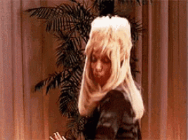 a woman in a blonde wig is standing in front of a palm tree .