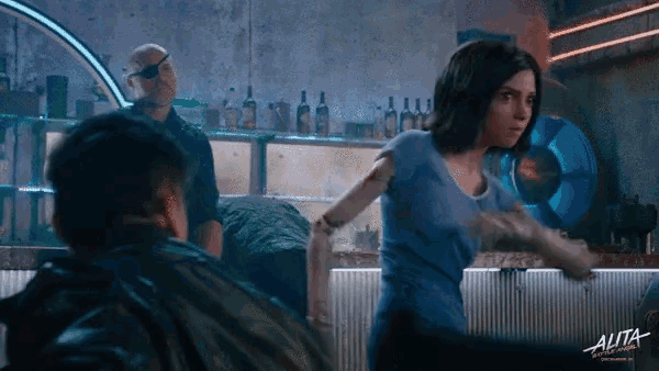 a woman in a blue shirt stands in front of a sign that says alita