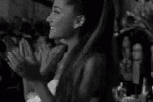 ariana grande is clapping her hands in a black and white photo while sitting in a crowd .