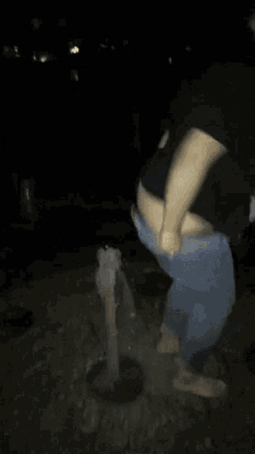 a man in a black shirt and blue jeans is squatting down in the dark while a woman stands behind him .