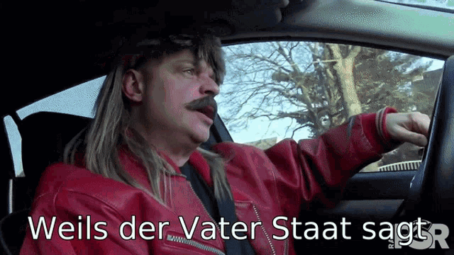 a man in a red jacket is driving a car with the words " weils der vater staat sagt " below him