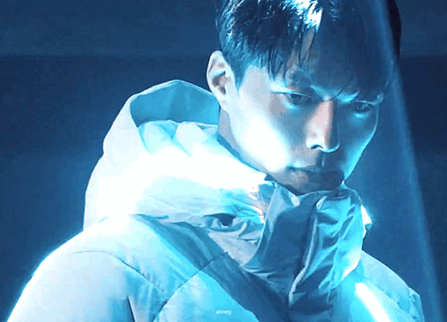 a man wearing a white jacket with a hood looks at the camera in a dark room