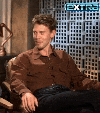 a man in a brown shirt sits in a chair in front of a screen that says extra
