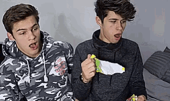 two young men are sitting next to each other on a couch and eating chips .