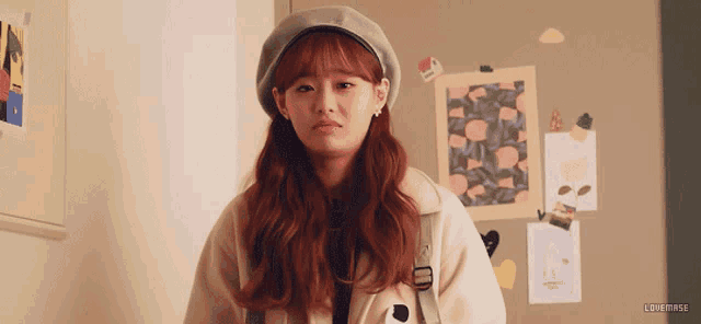 a girl with red hair wearing a beret and a white coat is standing in front of a wall with stickers on it