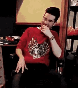 a man wearing a red sweater with a wolf on it is sitting in a chair and laughing .